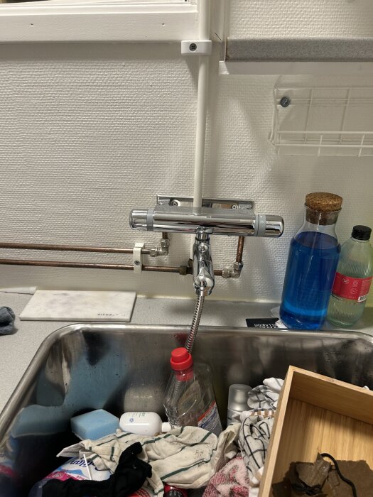 Vask i en tvättstuga med diverse rengöringsartiklar och tygstycken omkring, samt synlig vattenkran och flaskor med rengöringsmedel.