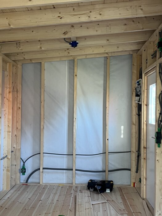 Inuti en oisolerad friggebod med synliga träreglar och elkablar dragna längs väggarna. En verktygslåda står på trägolvet.