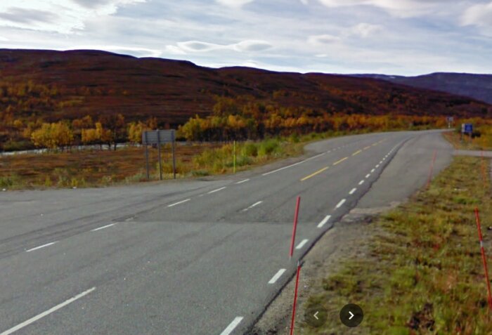 Landsväg med gul mittlinje i höstlandskap, vägskyltar på avstånd, indikerar gränsövergång mellan länder.
