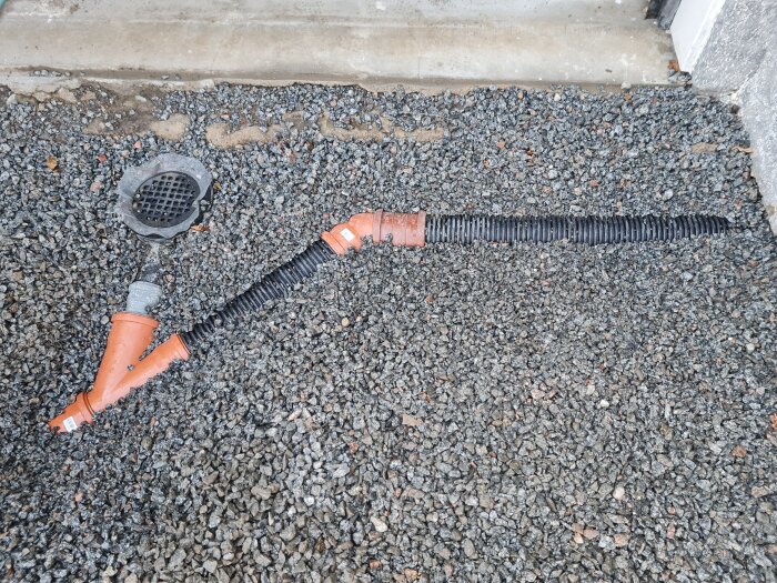 Orange och svarta dräneringsrör vid markytan, delvis täckta av grus, nära garageinfart med synlig betongkant.