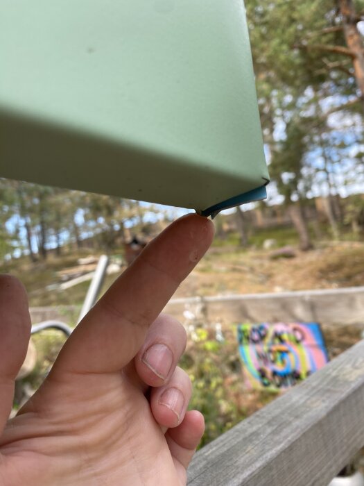 Finger pekar på vatten som droppar från hörnet av ett plåttak, omgiven av träd och en färgglad skylt i bakgrunden.