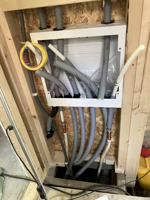 Kablage och rörinstallation i trävägg med oskyddade rör och kablar, flera grå slangar oordnade.