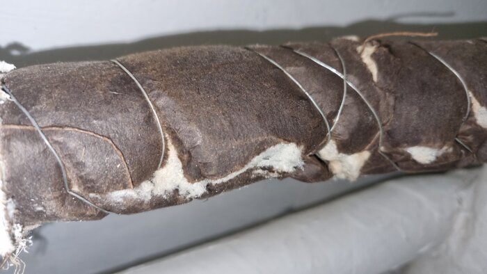 Brunt isoleringsmaterial runt ett rör, inlindat med metalltråd, med vita fibrer synliga vid sprickor.