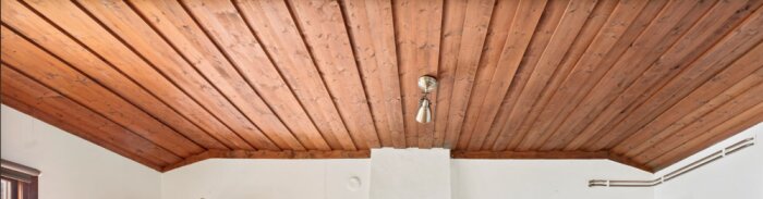 Innertak i vardagsrum med mörklackade träpaneler och centrerad taklampa. Vinklade sidopaneler syns, möjlig modernisering diskuteras.