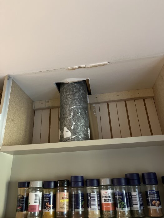 Ventilationsrör i kök, synligt genom en öppning i taket ovanför en hylla med kryddburkar.