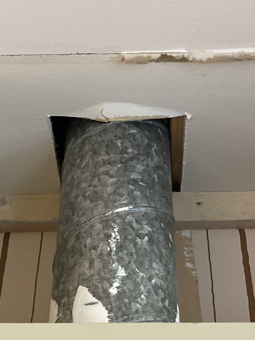 Ventilationsrör genom ett hål i ett gipstak med sprickor och skador runt hålet.