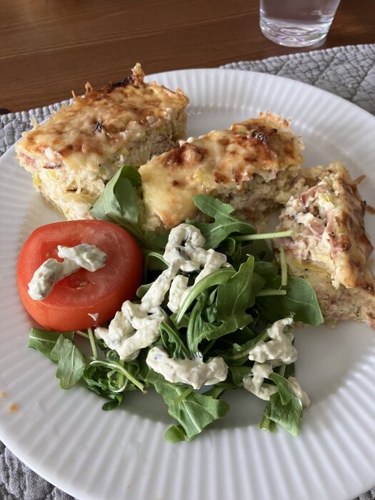 Kesogratäng med purjolök och skinka serverad med tomat och ruccola på vit tallrik.