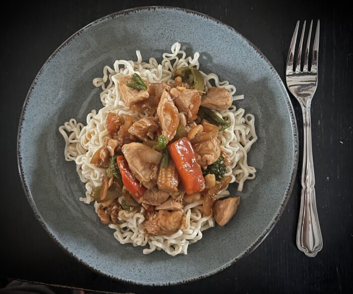 Wokad kyckling med grönsaker och jordnötter, serverat på en bädd av nudlar på en grå tallrik med en gaffel bredvid.