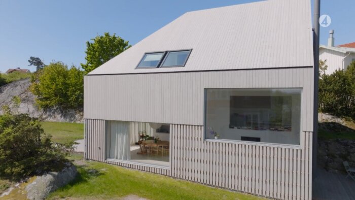 Modernt hus med sluttande tak, stora fönster och träribbad fasad, omgivet av grönska.