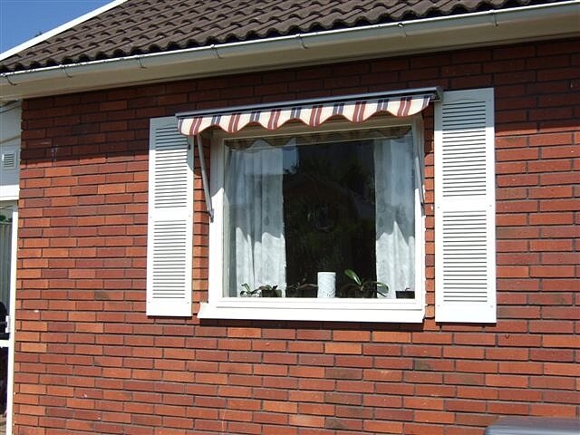 Fönster med randig markis och vita persienner på röd tegelvägg, del av husfasad.