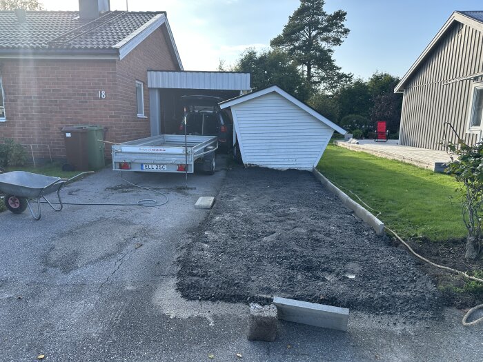 Bil och släp med krossmaterial på en uppfart, jord utlagd bredvid, skottkärra syns i förgrunden, och ett litet vitt skjul till höger.