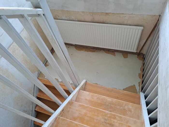 Trappa med furusteg och vitmålat räcke. Vilplan med golvspånskiva vid en vit radiator, omgiven av delvis borttagen tapet.