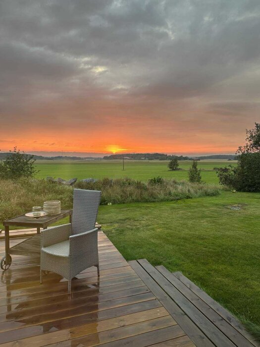 Solnedgång över fält från en träveranda med en stol och ett bord med dryck i förgrunden, molnig himmel i bakgrunden.