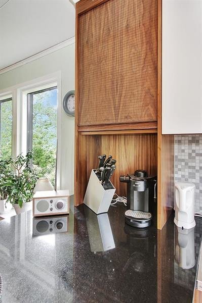 Modernt kök med svart bänkskiva, knivblock, kaffemaskin och högtalare. Fönster med grönska utanför och väggklocka synlig.