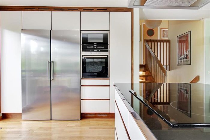 Modernt kök med rostfri kyl, integrerad ugn, induktionshäll, trägolv och öppen trappa i bakgrunden.