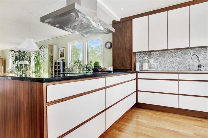 Modernt kök med vita skåp, trädetaljer, köksö, och rostfri fläktkåpa. Ljusinsläpp från fönster och inredning med växter och hushållsapparater.