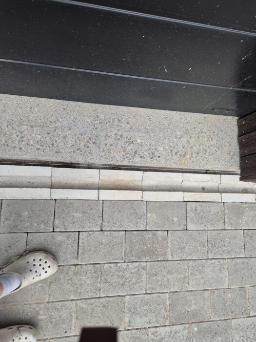 Ojämn stenläggning nära en vägg, där ett mellanrum syns mellan stenarna och väggen. En person står i bildens nedre vänstra hörn.