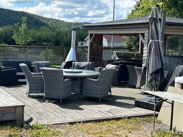 Utomhus veranda med utemöbler och öppen spis, omgiven av träd och buskar, en grillplats i bakgrunden och ett bord med stolar i fokus.