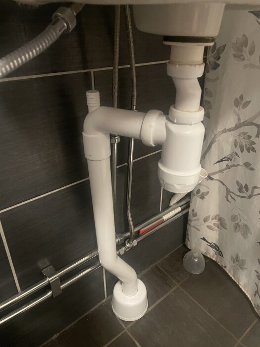 Vitt avloppssystem under handfat med möjlighet för tvättmaskinsanslutning efter vattenlås, synligt mot mörka kakelväggar.