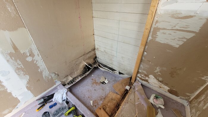 Fuktskadat hörn i badrum med svart mögel på gips och råspont. Verktyg och byggmaterial på golvet.
