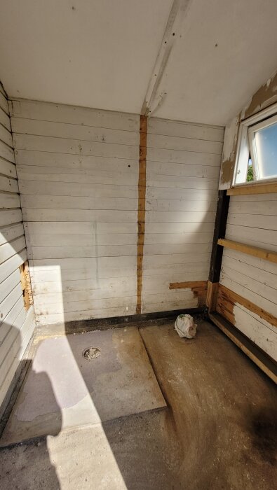 Vatten- och fuktskadade väggar i ett badrum under renovering, synliga träpaneler, cementgolv och ett fönster, med påsar i hörnet.