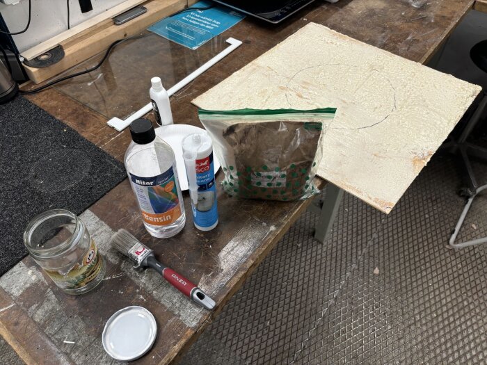 Material och verktyg för korklimningstest inklusive bensin, silikon, pensel, och pigment på en arbetsbänk.