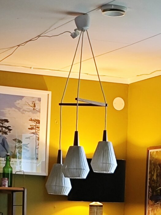 Taklampa med fyra vita lampskärmar som hänger i vitt tak, omgivna av ljusgul vägg och konst på väggarna.