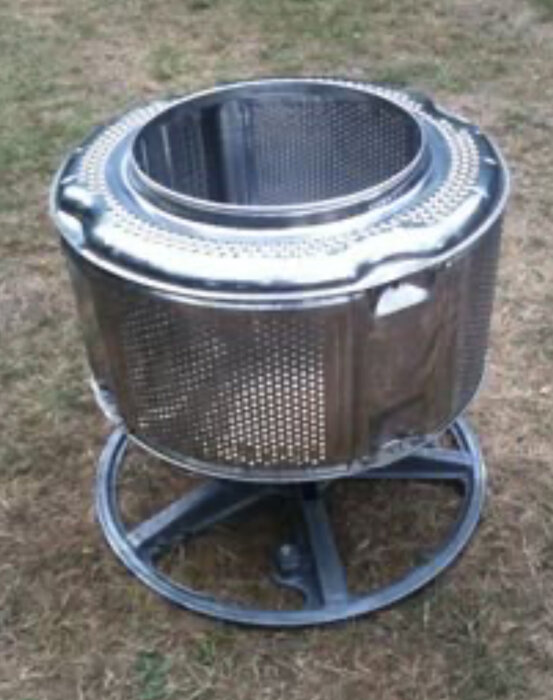 Gammal tvättmaskinstrumma används som eldstad, placerad på stativ på gräsmatta.