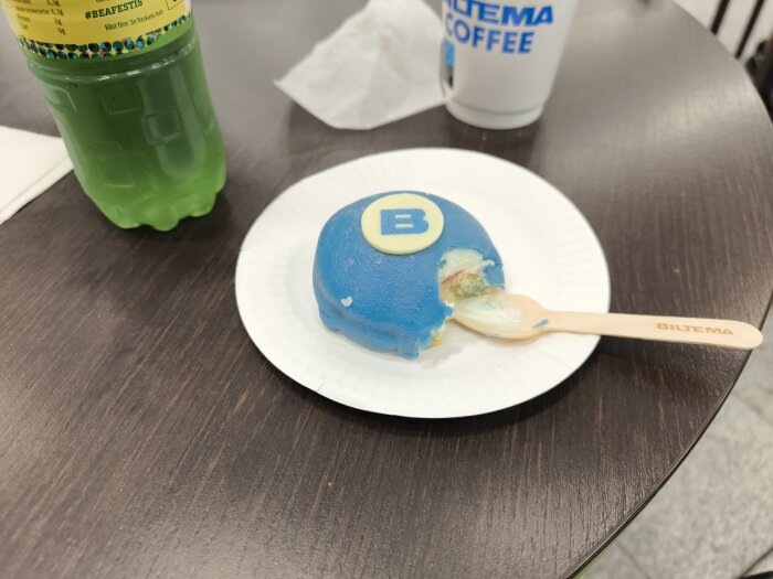 Blå bakelse med bokstaven "B" på, upplagd på vit papptallrik med en träsked bredvid. Grön dryck och Bitr-ma kaffe i bakgrunden.
