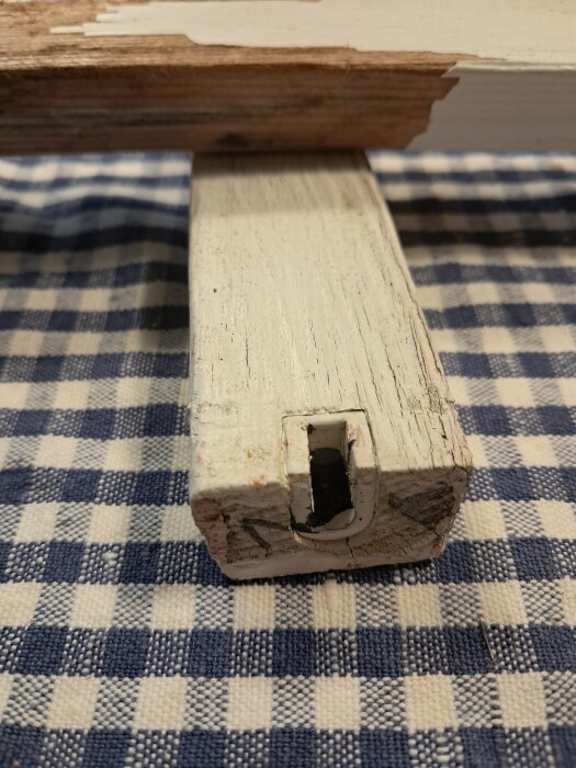 Gammalt, vitmålat spröjsfäste i trä med slitage, liggande på blå- och vitrutig duk.