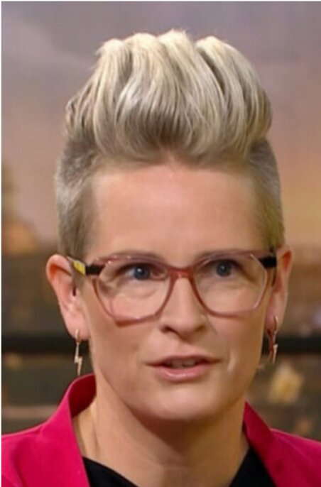 En person med stilfull, uppåtstående frisyr, glasögon, rosa kavaj och örhänge i form av blixt.