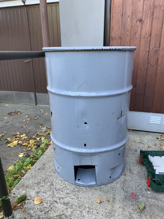 Metallfat med flera hål och en flärp som sticker ut nedtill, placerad utomhus på en betongyta nära en byggnad.