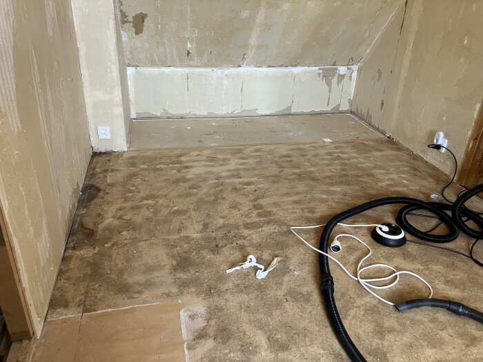 Masonitskivor med lim och luddrester från gamla mattor på golv, sladdar och en dammsugare syns i vänstra hörnet av det tomma rummet.