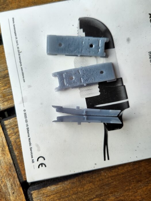 Tre grå 3D-printade reservdelar till ett larm ligger på ett papper med text och svartvit grafik.