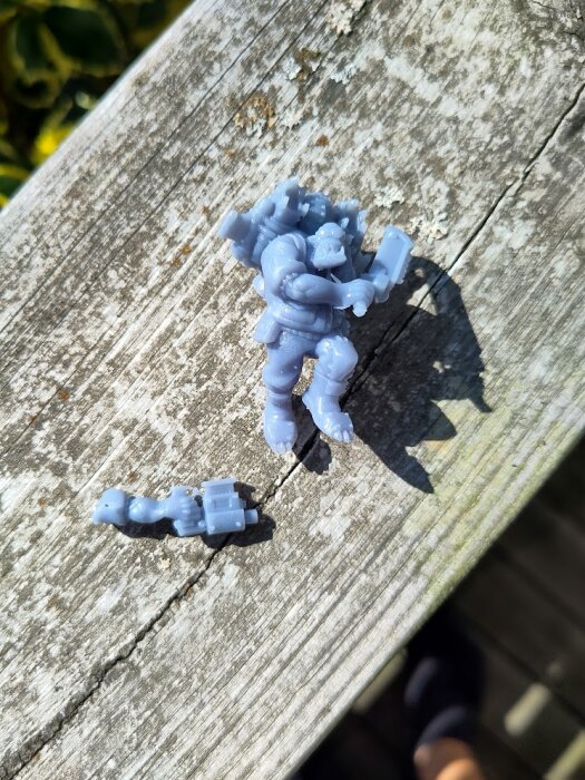 3D-printad grå minifigur i rustning med en löstagbar del, placerad på en träyta utomhus.