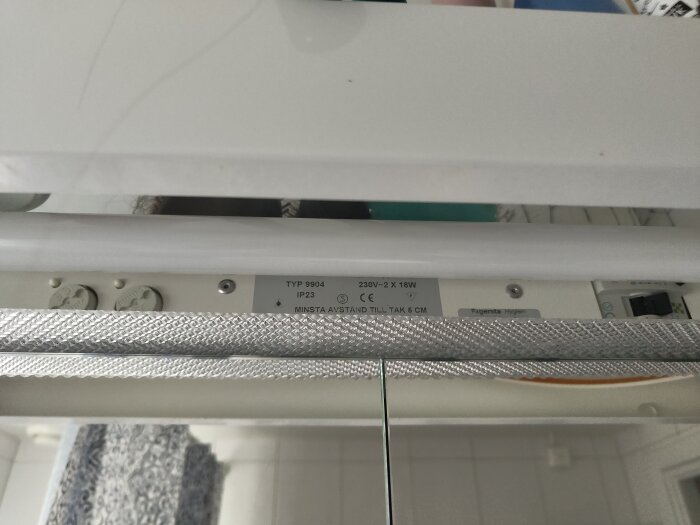 Närbild av en lysarmatur med texten "TYP 9904 IP23" och en switch ovanpå. Armaturen sitter nära en dusch med duschdraperi.