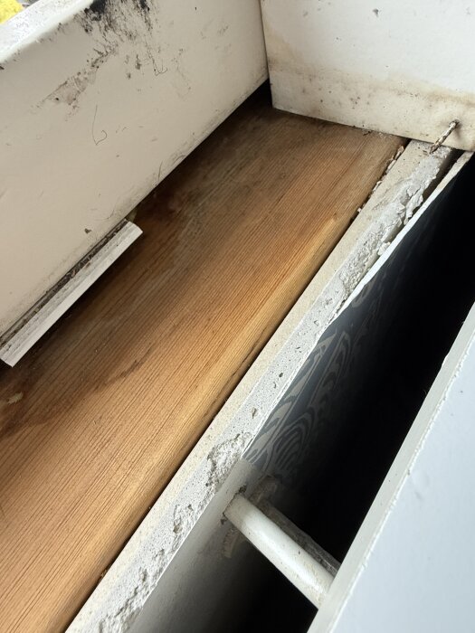 Blött hörn på trä, exponerat underkant av fönster med avlägsnad smygbräda; tecken på fuktskada i drevet bakom, vit mur, rör synligt.
