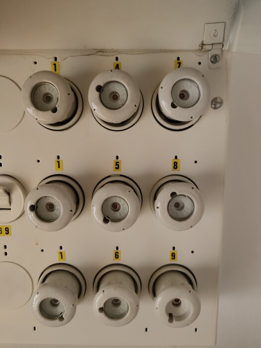 Proppskåp med nio äldre porslinsproppar, var och en numrerad med klisterlappar, monterat på en vit vägg.