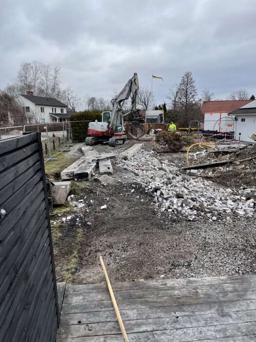 Grävmaskin på rivningsplats med spillror av ett nerbrunnet hus i bakgrunden, omgiven av staket och en flagga som vajar i vinden.