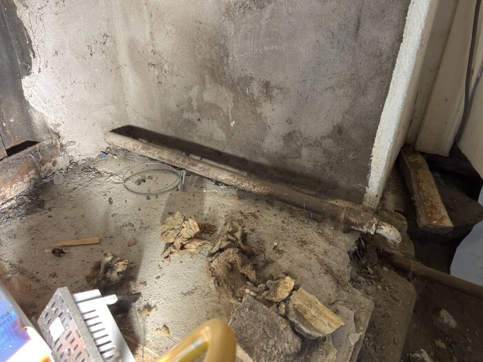 Ett rostigt rör på en betonggolv med skräp runt, bredvid en vägg i renoveringsmiljö.