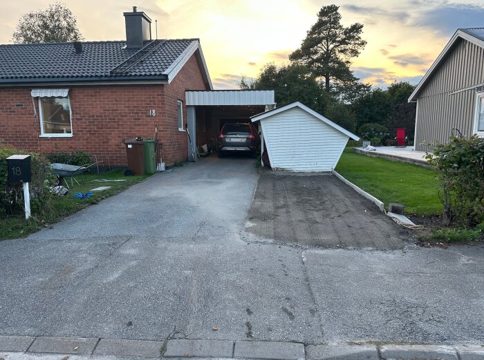 Uppfart framför ett rött tegelhus med en bil i garaget. Delvis asfalterad och grusig yta med nya kantstenar. Gräsyta och buskar till höger.