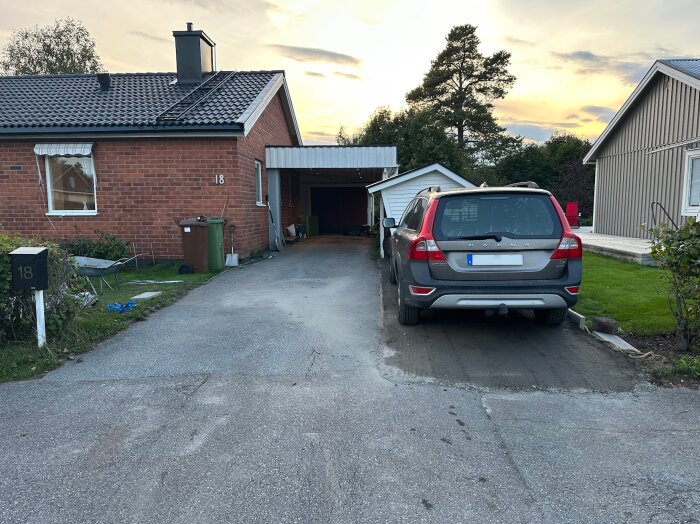 Bil parkerad på en uppfart vid ett rött tegelhus med carport och garage. Kantstenar syns vid sidan och en grön gräsmatta till höger.