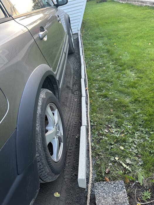Grå bil parkerad bredvid kantstenar som inte är helt nedgrävda i marken intill en gräsmatta. Kantstenarna lutar och är inte stabila.