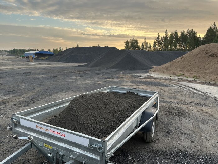 Släpvagn med mörkt grus framför sand- och grushögar vid stenbrott i gryningen.