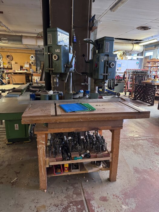 Två pelarborrar av märket Arboga 2512 i verkstad nära arbetsbord fullt med diverse borrar och tillbehör.