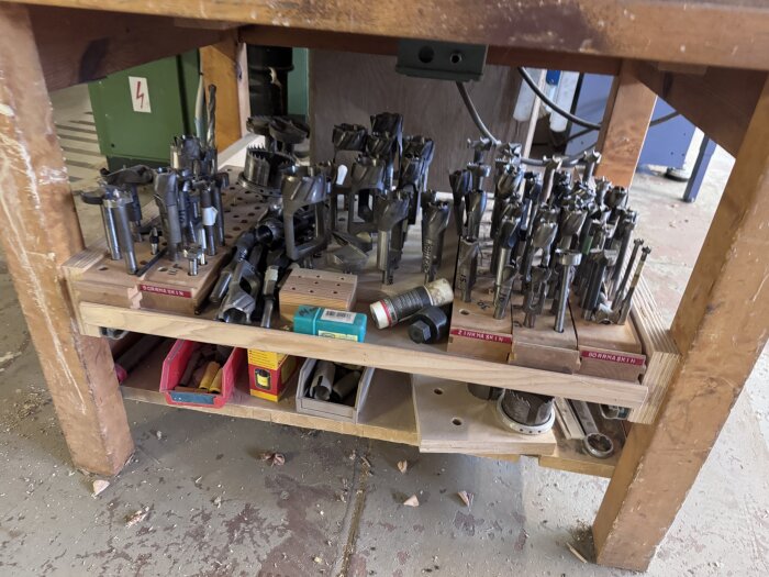 Flera borrhuvuden och tillbehör organiserade på en hylla under ett träbord; etiketter visar texten "Zinkmaskin" och "Borrmaskin".