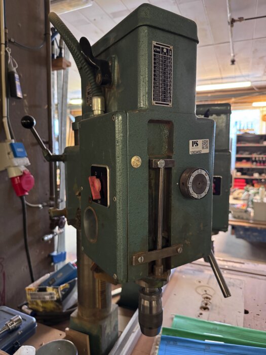 Grön pelarborrmaskin Arboga 2512 med reglage och graderad ratt, omgiven av verktyg och tillbehör i ett garage eller verkstad.