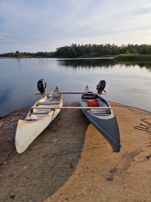 Två kanoter med motorer, förenade av en träbalk, ligger på en betongbrygga vid sjö. Tittar mot vatten och skog i bakgrunden.