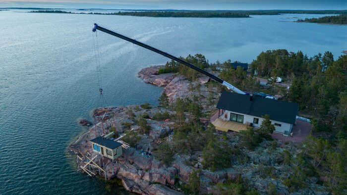 Mobilkran lyfter strandbastu på plats vid kusten, omgivet av skog och klippor.