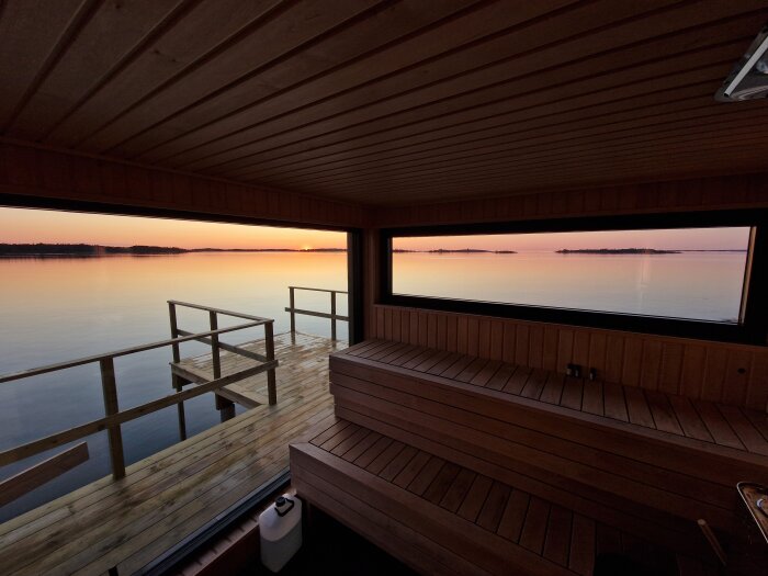 Vy från en bastu med stora fönster som vetter ut mot en sjö, där solnedgången färgar himlen och vattnet i varma toner.